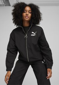 Sort zip-jakke lavet af letvægtsstof, med et hvidt Puma-logo på brystet og kontrasterende hvide striber langs skuldrene.