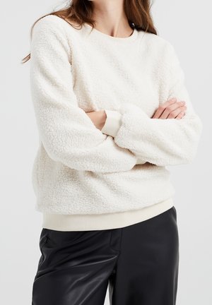 Kvinde iført en hvid tekstureret fleece-trøje med lange ærmer og sorte læderbukser, arme krydset.