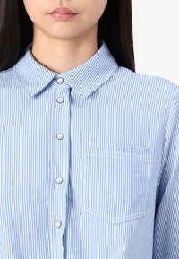 Blue and white striped button-up shirt with a folded collar, featuring a chest pocket and silver buttons along the front.