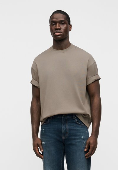 Άνδρας που φοράει χαλαρό ταυπί t-shirt με ρεβέρ στα μανίκια και σκούρα μπλε τζιν με φθορές, στέκεται μπροστά σε ένα απλό φωτεινό φόντο.