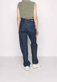 Jeans de mezclilla de color azul oscuro con cintura alta, corte recto y costuras visibles. Logo en la cinturilla negra. El modelo lleva zapatillas blancas.