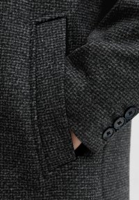 Grå tekstureret frakke med en lomme, tre-knaps mansjet med sorte knapper og et diskret ternet mønster.