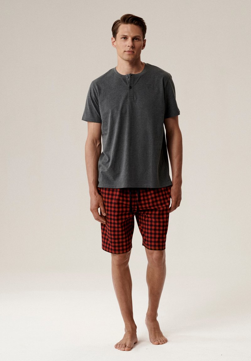 Szary, krótki t-shirt henley z trzema guzikami, sparowany z czerwonymi krótkimi spodniami w kratę. Materiał wydaje się miękki i ma luźny krój.