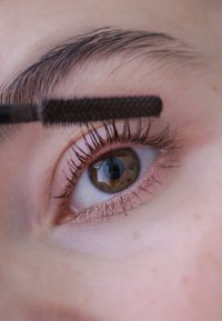 Primer plano de un ojo con largas pestañas rizadas y una ceja oscura. Un aplicador de máscara, con un cepillo texturizado, está posicionado sobre las pestañas.