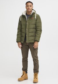Puffjakke i olivengrøn, med hætte, stort quiltet design, pelskrave og brune knapper. Sammen med cargo-bukser og brune støvler.