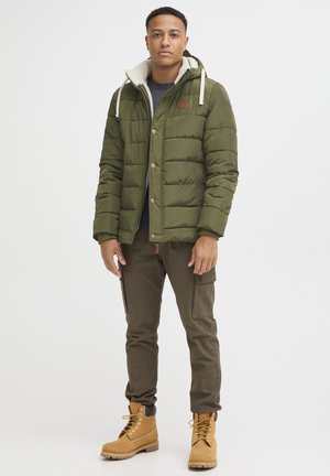 Chaqueta acolchada verde oliva con cuello de piel sintética, botones beige y parche con logo, combinada con pantalones cargo oscuros y botas color canela.