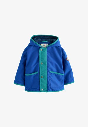 Lille barn står iført en blå og grøn jakke, lyse vidde jeans og navy sneakers med grønne striber på en lys baggrund.