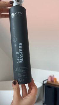 Une main tient un flacon noir de 500 ml de laque Revlon Style Masters à tenue forte dans une salle de bain avec une baignoire et un chariot en arrière-plan.