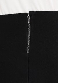 Svart stickad plagg med silverfärgad dragkedja. Har en kontrasterande vit panel upptill. Slät textur, figurnära design.