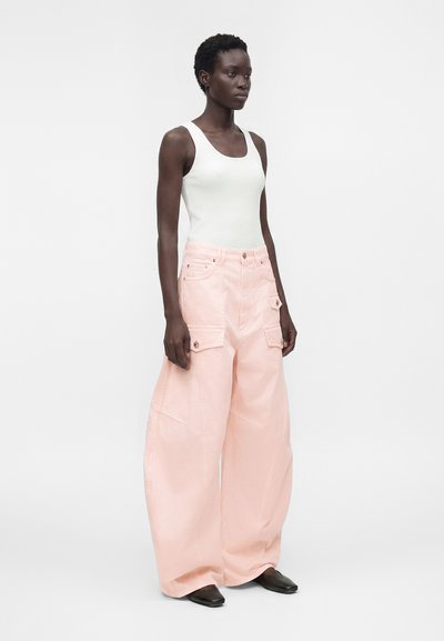 Γυναίκα όρθια, φορώντας λευκό αμάνικο ριμπ top, φαρδιά ανοιχτό ροζ cargo παντελόνια με τσέπες και μαύρα μυτερά παπούτσια, ουδέτερο φόντο.