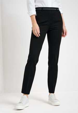 Frau trägt schwarze Slim-Fit-Hose, weiße Sneakers und ein weißes Hemd und steht vor einem einfarbigen hellen Hintergrund.