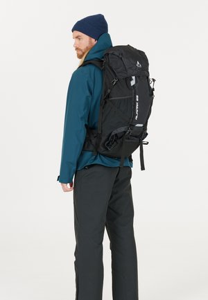 Zwart wandelrugzak met meerdere banden, gaas zijvakken en een bovenklep, gedragen door een persoon in een teal jack en zwarte broek.
