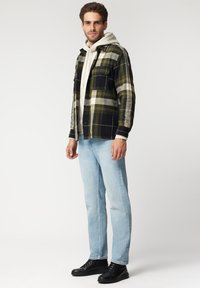 Czarna, zielona i kremowa koszula w kratę z flaneli z kieszeniami zapinanymi na guziki, nałożona na szary hoodie, w połączeniu z jasnoniebieskimi dżinsami i czarnymi sneakersami.