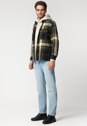 Czarna, zielona i kremowa koszula w kratę z flaneli z kieszeniami zapinanymi na guziki, nałożona na szary hoodie, w połączeniu z jasnoniebieskimi dżinsami i czarnymi sneakersami.