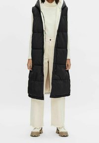 Doudoune noire sans manches avec capuche, présentant un design matelassé et une fermeture éclair. Portée sur un pull crème et un pantalon large de couleur claire.