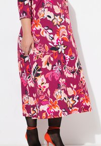 Vestido floral en fucsia con patrones en naranja, rosa y negro. Confeccionado con mangas de tres cuartos y corte hasta la rodilla. Combinado con tacones naranjas.