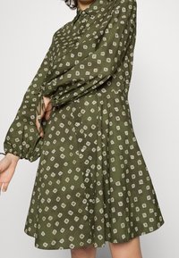 Abito verde con scollo a V e motivo a diamante, maniche lunghe con volume e dettagli a fiocco ai polsini. Tessuto leggero con una forma svasata.