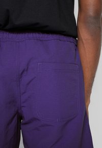 Violetit housut, joissa on teksturoitu kangas, joustava vyötärönauha ja takatasku, jossa on kontrastivärinen ommel. Design on yksinkertainen ja toimiva.