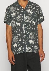 Camicia a maniche corte in tessuto grigio scuro con una stampa bianca che ricopre tutta la superficie, raffigurante animali, forme geometriche e simboli celesti. Bottoni frontali visibili.