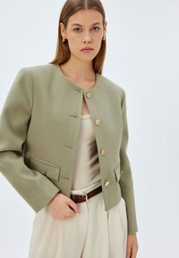Hellgrüne, strukturierte Jacke mit rundem Halsausschnitt und goldenen Knöpfen. Hat Fronttaschen und lange Ärmel. Kombiniert mit einem beigen Oberteil und weißen Hosen.