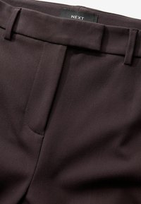 Sötétbarna, testre szabott nadrág sima szövettextúrával, egy elülső zsebbel és tűzött derékpánttal. Márkacímke látható.