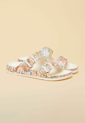 Paar weiße Slides-Sandalen mit mehrfarbigem Blumenmuster und zwei verstellbaren Schnallenriemen auf beigem Hintergrund.