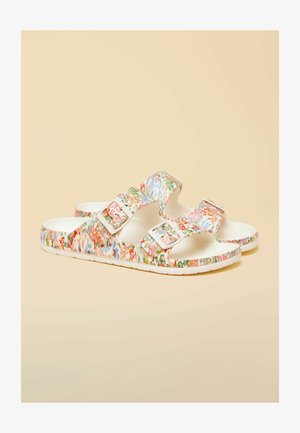 Par hvide slidssandaler med flerfarvet blomstermønster og to justerbare stropper med spænde på beige baggrund.