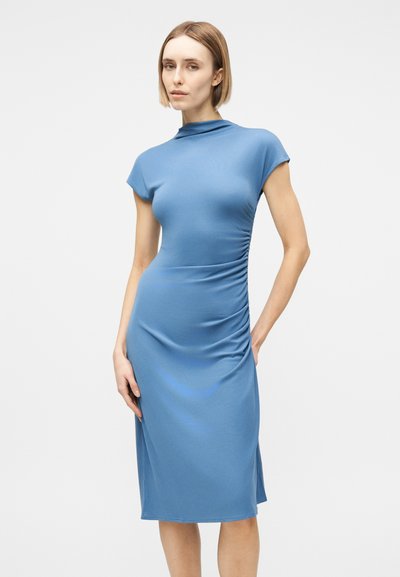 Γυναίκα που στέκεται, φορώντας ένα εφαρμοστό μπλε φόρεμα μήκους έως τα γόνατα με κοντά μανίκια και ελαφριά ρίγα στη μία πλευρά, μπροστά σε απλό λευκό φόντο.