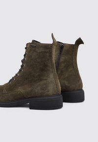 Botas de tobillo de ante verde oliva con cordones en la parte delantera, cierre con cremallera en la parte trasera y suelas de goma negras. Cuenta con una lengüeta para facilitar su uso.