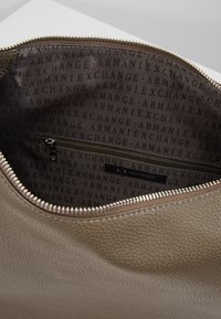 Interno della borsa beige con texture, dotato di tasca con zip e stampa del logo in marrone scuro. Zip liscia con accento metallico.