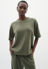 Žena s krátkými vlasy, oblečená v olivově zelené volné košili s krátkým rukávem a odpovídajících kalhotách, stojící s rukama v kapsách před jednoduchým pozadím.