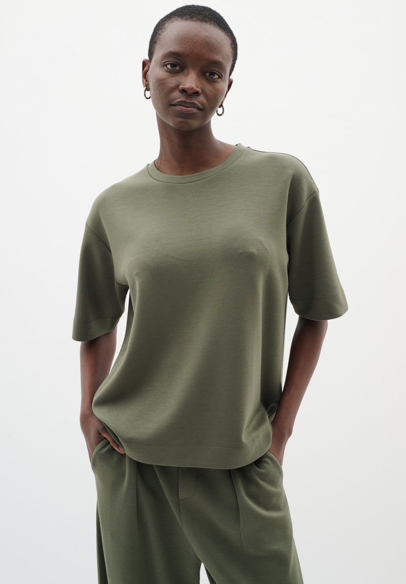 Žena s krátkými vlasy, oblečená v olivově zelené volné košili s krátkým rukávem a odpovídajících kalhotách, stojící s rukama v kapsách před jednoduchým pozadím.