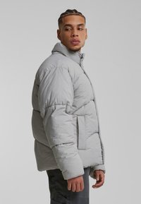 Chaqueta de plumas de color gris claro con aislamiento grueso, cuello alto y bolsillos frontales. Textura suave y forma cuadrada para calidez y comodidad.