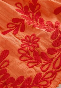 Orange fabric with raised red floral and leaf embroidery detail, showing texture and stitching patterns.