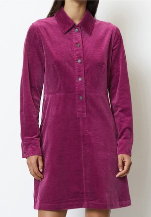 Femme portant une robe en velours côtelé magenta à manches longues, boutonnée et avec col, debout devant un fond blanc uni.