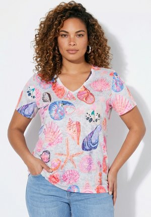 Kvinde med krøllet hår iført store øreringe og en hvid V-hals T-shirt med farverige motiver af muslinger og søstjerner, kombineret med lyseblå jeans.