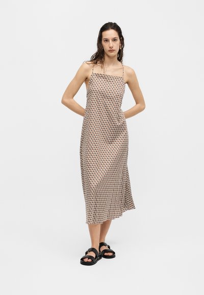 Γυναίκα φοράει μίντι φόρεμα χωρίς μανίκια με γεωμετρικό σχέδιο, λεπτές τιράντες και μαύρα σανδάλια, στέκεται μπροστά σε απλό λευκό φόντο.