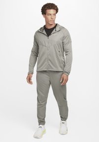 Chaqueta atlética gris claro con capucha, cierre frontal y paneles texturizados, combinada con pantalones a juego y zapatillas blancas con acentos amarillos.