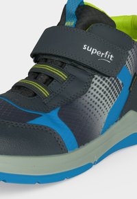 Baskets enfant bleu et gris avec bride velcro étiquetée "superfit,", lacets élastiques, accents vert néon et semelle texturée.