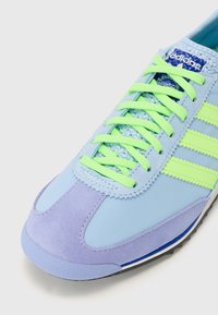 Zapatilla azul claro con acentos de ante morado, cordones neón verdes y tres rayas verdes. Parte superior texturizada con lengüeta de malla y suela de goma.