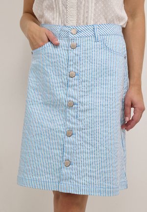 Femme portant une jupe boutonnée à rayures verticales bleues et blanches, main dans la poche, associée à un chemisier sans manches blanc texturé.