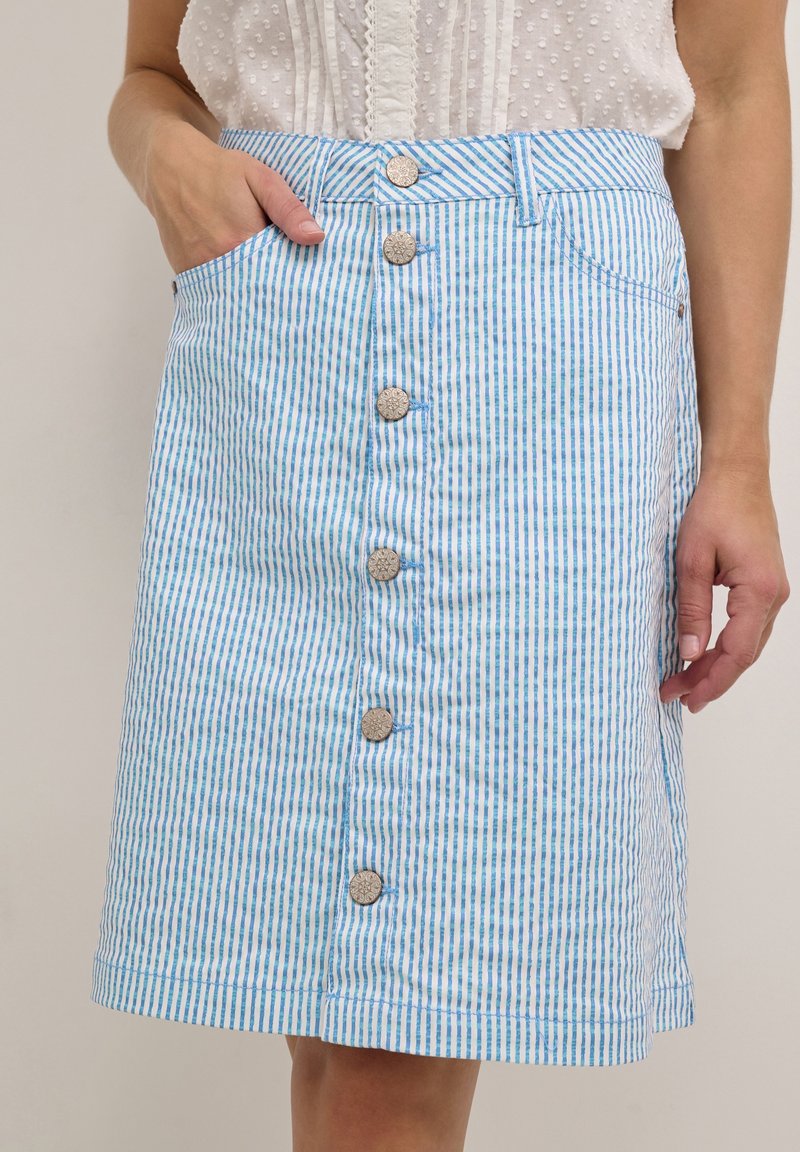 Femme portant une jupe boutonnée à rayures verticales bleues et blanches, main dans la poche, associée à un chemisier sans manches blanc texturé.
