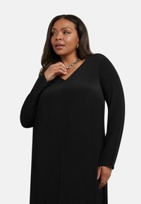 Svart långärmad klänning med V-ringning, slät textur och sidofalsar. Ett silverhalsband ger en metallisk accent.