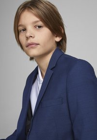 Blazer bleu marine en tissu texturé, avec une poche poitrine simple et des revers crantés, porté par-dessus une chemise à col blanc.