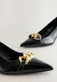 Le décolleté neri a punta abbinate presentano un dettaglio decorativo con catena dorata sul davanti, su uno sfondo neutro.