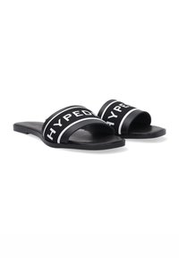 Zwarte slip-ons met brede stoffen banden met witte letters, platte rubberen zolen en een minimalistische constructie.
