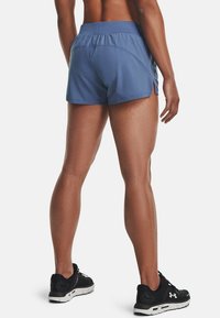 Person med blå träningsshorts och svarta löparskor, stående med ryggen mot kameran mot en vit bakgrund.