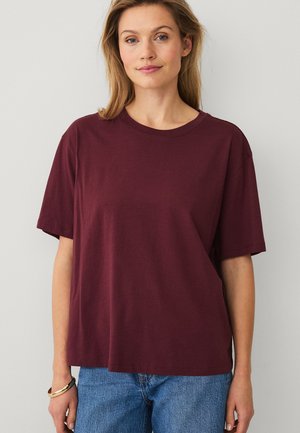 Woman wearing a loose-fitting maroon short-sleeve t-shirt and blue jeans, standing against a plain white wall.