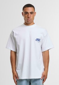 Valkoinen puuvillapaita, jossa on pyöreä kaula-aukko, lyhyet hihat ja sininen painatus vasemmassa rinnassa. Normaali istuvuus ja sileä pinta.