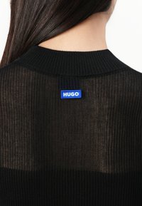 Mustan neulepaidat, joissa on uritettu pinta ja klassinen kaula-aukko. Takana on sininen suorakaiteen muotoinen etiketti, jossa lukee "HUGO".